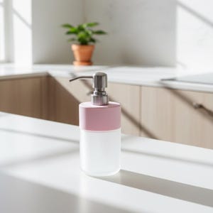 Op de afbeelding: Een moderne zeepdispenser met een basis van matglas en een lichtroze bovenkant. De dispenser heeft een zilveren pomp en staat op een wit aanrechtblad. Een kleine plant in een pot staat op de achtergrond.