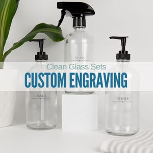 May include: Three clear glass bottles with black pump tops and labels. The labels read "GLASS", "HANDS", and "DISHES". The bottles are on a white surface with the text "Clean Glass Sets CUSTOM ENGRAVING" in blue.