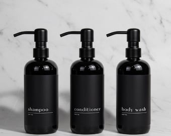 May include: Three black cylindrical plastic bottles with matte black pump dispensers. Each bottle features a black label with white text reading shampoo, conditioner, or body wash, and the capacity 500 ml printed below the text in a clean font.