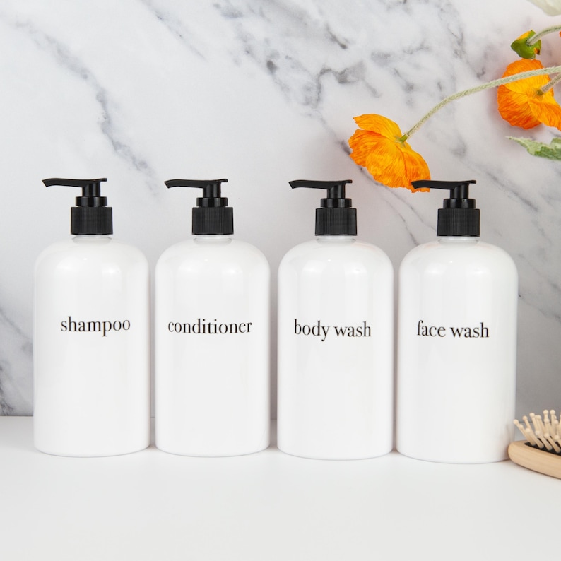 May include: Four white pump bottles labelled "shampoo", "conditioner", "body wash", and "face wash". Each bottle has a black pump top. The bottles are in a row, with a marble background and orange flowers.