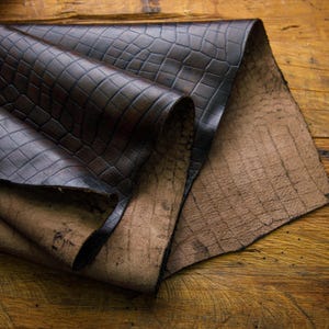 May include: Two pieces of brown leather with a crocodile-embossed pattern. The top piece is folded over the bottom piece.