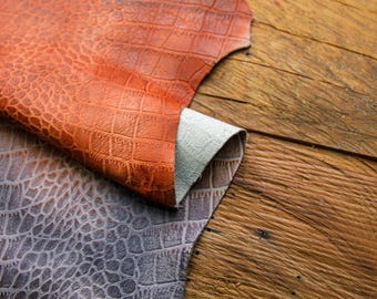 May include: Two pieces of leather with a crocodile pattern. One piece is orange and the other is gray. The leather is lying on a wooden surface.