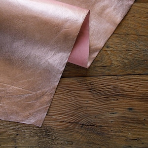 May include: A piece of rose gold leather folded over on a wooden surface. The leather has a metallic sheen and a textured surface.