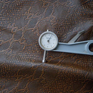 May include: A close-up of a brown faux leather fabric with a snake skin pattern. A dial gauge is placed on top of the fabric. The dial gauge reads "0.1 mm" and is labeled "GALVAN" with a "Made in Germany" inscription.