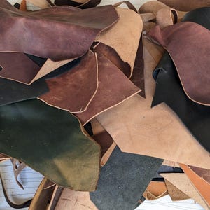 May include: A pile of leather scraps in various shades of brown, black, and green. The leather is cut into different shapes and sizes, and some pieces have a distressed or aged look.