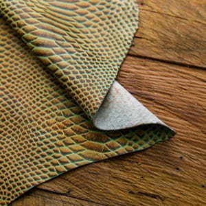 May include: A piece of green and brown snakeskin leather with a textured pattern. The leather is folded over on itself and is lying on a wooden surface.