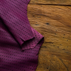 May include: A close-up of a piece of purple leather with a diamond-shaped hole pattern. The leather is folded over on itself and is lying on a wooden surface.