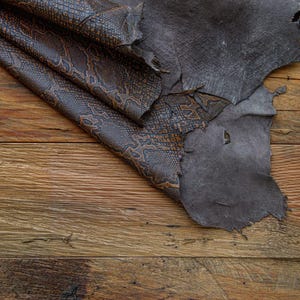 May include: A piece of brown leather with a snake skin pattern is folded over a piece of dark gray leather. Both pieces of leather are on a wooden surface.