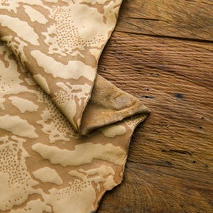 May include: A close-up of a beige leather fabric with a camouflage pattern. The fabric is folded and has a rough texture. The fabric is on a wooden surface.