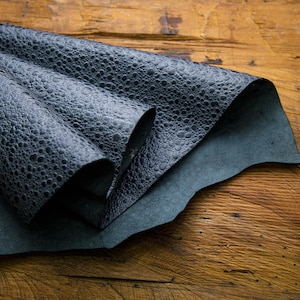 Può includere: Primo piano di pelle nera con una superficie testurizzata e ghiaiosa. La pelle è piegata in tre parti, rivelando una pelle liscia blu scuro sottostante.