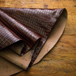 May include: Two pieces of brown leather with a crocodile pattern, folded and stacked on top of each other. The leather is textured and has a glossy finish.