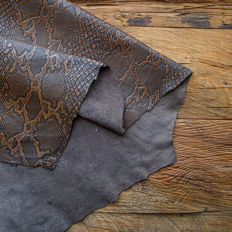 May include: A piece of brown leather with a snakeskin pattern folded over a piece of gray leather. The leather is on a wooden surface.