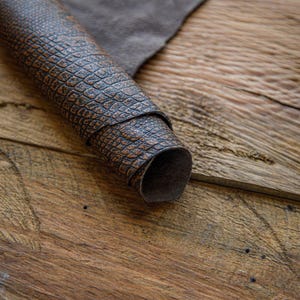 May include: A close-up of a brown leather with a textured, alligator-like pattern. The leather is rolled up and lying on a wooden surface.