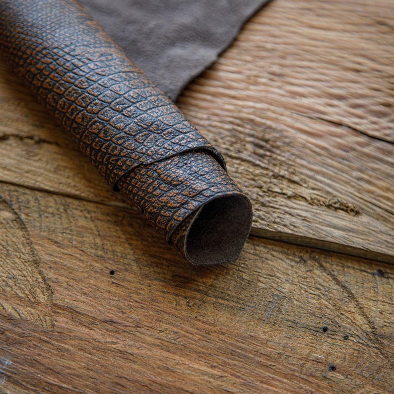 May include: A close-up of a brown leather with a textured, alligator-like pattern. The leather is rolled up and lying on a wooden surface.