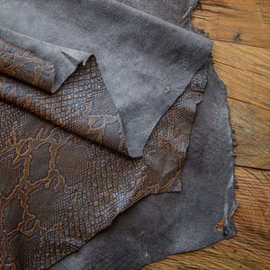 May include: Two pieces of leather, one brown with a snake skin pattern and one gray, are folded together on a wooden surface.