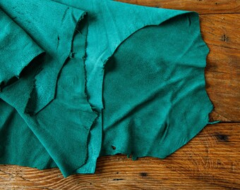 May include: A piece of teal-colored leather with a rough, uneven edge, lying on a rustic wooden surface. The leather is folded and appears soft, with visible texture and slight variations in color.