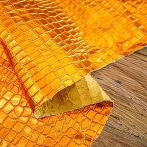 May include: A close-up of a gold-coloured fabric with a crocodile skin pattern. The fabric is folded and draped over a wooden surface.