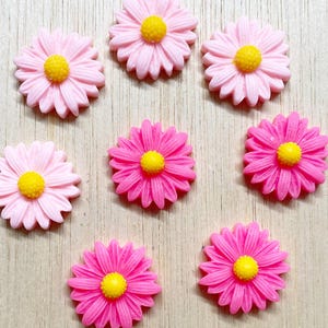 May include: Eight flower-shaped embellishments in various shades of pink with yellow centers, arranged in a group. The text 8 Piece pink mix is printed at the top. Each piece features a daisy-like design with multiple thin petals radiating from a center.