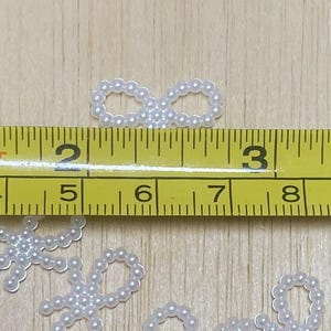 May include: White pearl embellishments in the shape of circles and infinity symbols. The pearls are approximately 1/2 inch in diameter. A ruler is included in the image for scale.