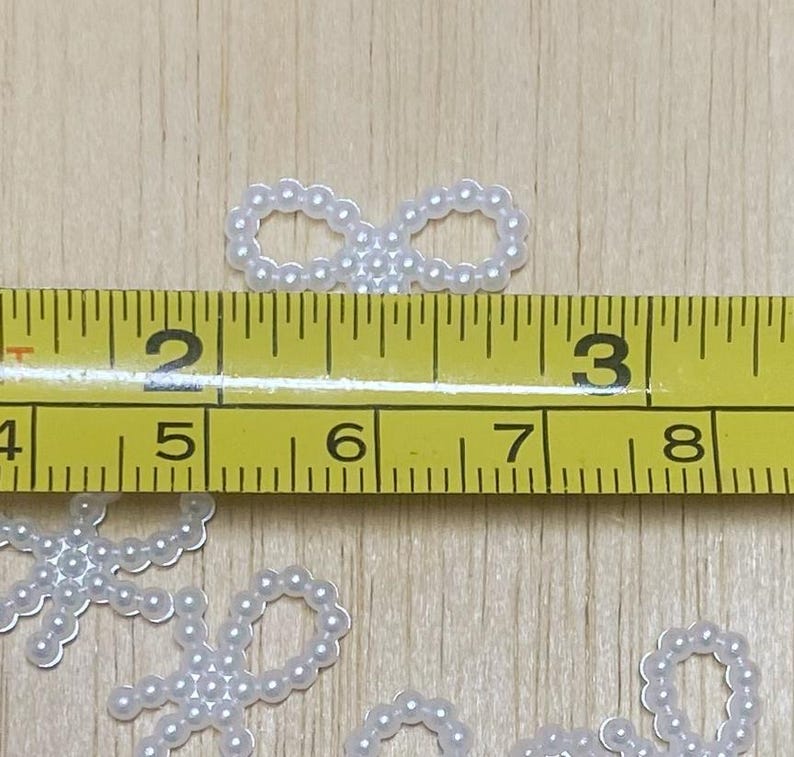 May include: White pearl embellishments in the shape of circles and infinity symbols. The pearls are approximately 1/2 inch in diameter. A ruler is included in the image for scale.