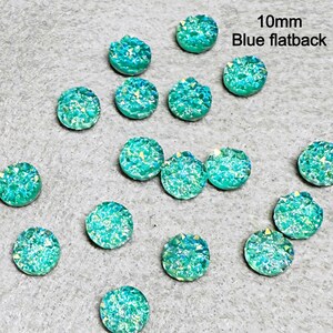 May include: A collection of 10mm blue flatback cabochons with a sparkling, textured surface. The cabochons are round and have a light blue color with iridescent accents. The text "10mm Blue flatback" is visible in the image.