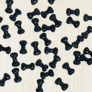 May include: A collection of small, black bow-tie shaped embellishments scattered across a light-colored surface. Each bow has a glittery texture, adding a touch of sparkle. These decorative elements are ideal for craft projects.