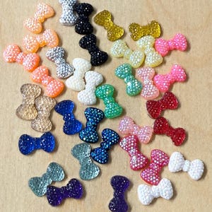 May include: A collection of small, bow-shaped embellishments in various colors, including orange, silver, black, gold, white, green, blue, pink, red, and purple. Each bow is covered in sparkling rhinestones, adding a touch of glamour.