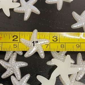May include: A collection of pearlescent white starfish, approximately 1 inch in size, are scattered across a dark surface. The starfish have a textured surface and are measured with a yellow measuring tape.
