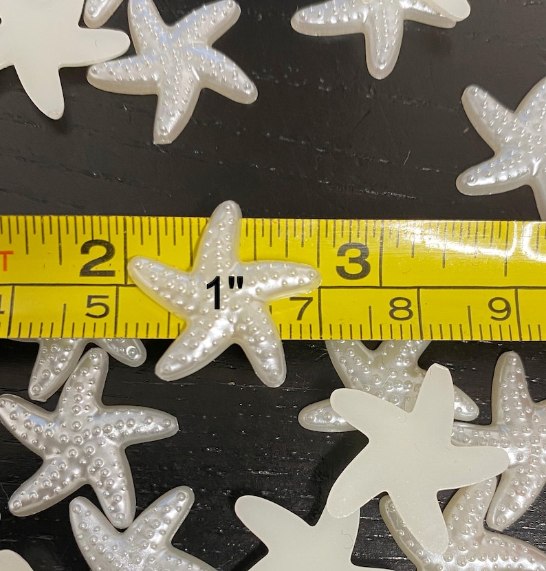 May include: A collection of pearlescent white starfish, approximately 1 inch in size, are scattered across a dark surface. The starfish have a textured surface and are measured with a yellow measuring tape.
