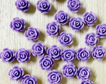 May include: A collection of small, light purple rose-shaped cabochons scattered across the frame. Each individual piece features a molded, multi-layered petal design with a smooth, glossy finish, creating a uniform appearance throughout the group.