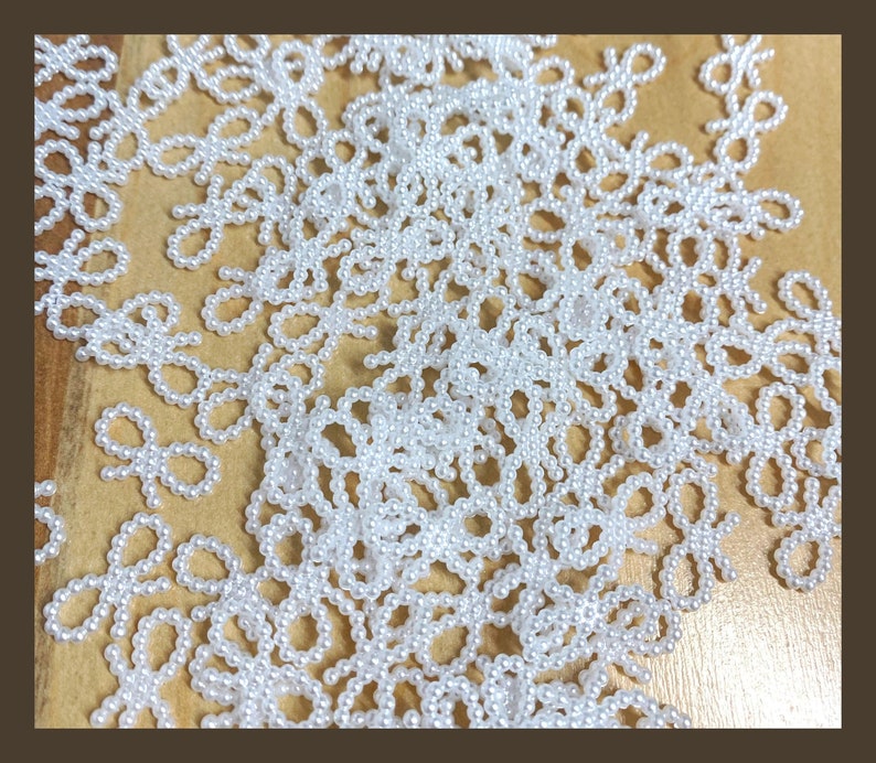 May include: A close-up of a large quantity of white pearl beads arranged in a pattern of bows. The beads are scattered on a brown surface.