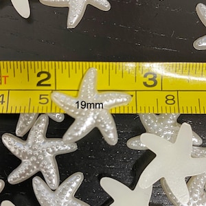 May include: A collection of pearlescent white starfish-shaped embellishments. The starfish are approximately 0.75 inches (19mm) in size. The image shows a yellow measuring tape next to the starfish.
