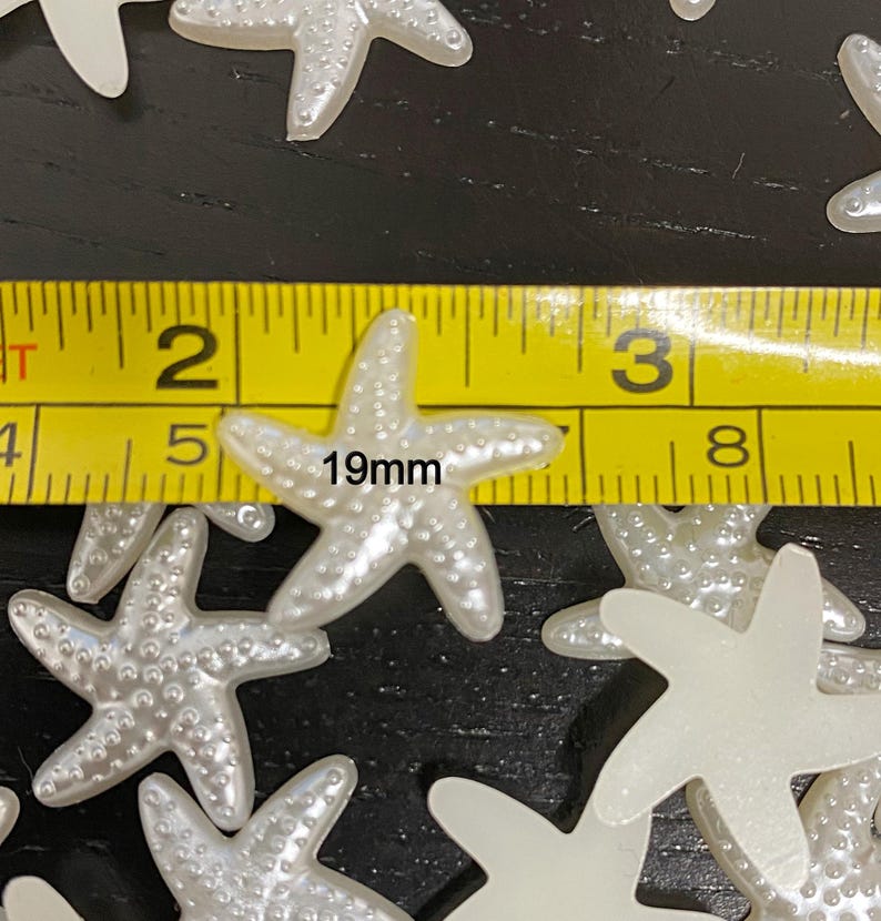 May include: A collection of pearlescent white starfish-shaped embellishments. The starfish are approximately 0.75 inches (19mm) in size. The image shows a yellow measuring tape next to the starfish.