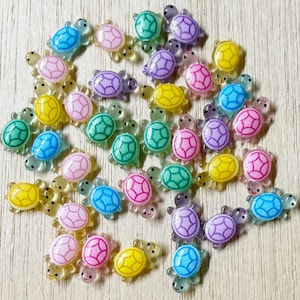 May include: A collection of small, translucent turtle-shaped cabochons in various colors including pink, blue, yellow, green, and purple. Each turtle features a distinct shell pattern with defined lines and a smooth, glossy finish on the surface.