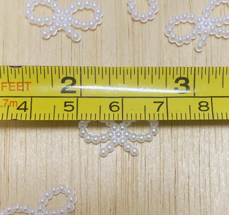 May include: White pearl bow embellishments on a wooden surface with a measuring tape showing 5 inches.