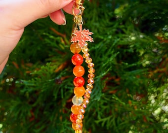 May include: A gold-toned metal keychain featuring a small red maple leaf charm attached to a gold lobster clasp. The keychain includes a vertical strand of smooth, round orange and yellow beads alongside a parallel strand of faceted amber crystals.