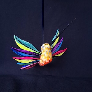 Puede incluir: Una colorida escultura de colibrí con un largo pico negro y alas multicolores. El cuerpo del ave es naranja y amarillo, y las alas son una mezcla de azul, verde, amarillo, morado y rosa. La escultura está suspendida sobre un fondo negro.