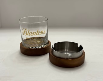 May include: A wooden coaster with a glass tumbler on top. The tumbler has the words "Blanton's The Original Single Barrel Bourbon" printed on it in gold lettering.  Next to the coaster is a wooden ashtray with a stainless steel insert.