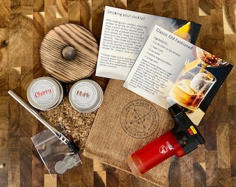May include: A wood smoking kit for cocktails, with two small tins of wood chips labeled "Cherry" and "Maple", a wooden lid, a small bag of wood chips, a metal straw, a small bag of metal mesh filters, a red and black torch lighter, and two recipe cards for a "Classic Old Fashioned" cocktail.
