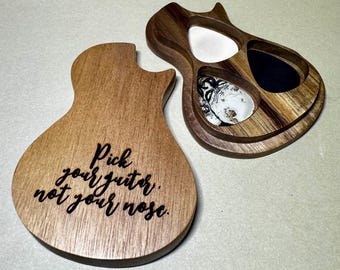 May include: Wooden guitar pick holder with a lid. The open holder contains guitar picks in white, black, and patterned designs. The lid has the phrase "Pick your guitar, not your nose" engraved in black.