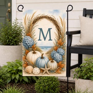 May include: A rectangular garden flag featuring a central letter M surrounded by a wreath of dried grasses, blue hydrangea flowers, and orange foliage. The scene includes three pumpkins, a seashell, and a starfish on a sandy beach background.