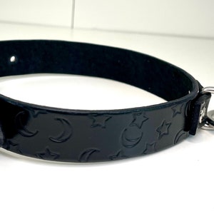 May include: Black leather collar with a moon and star pattern embossed on the surface. The collar has two silver D-rings.