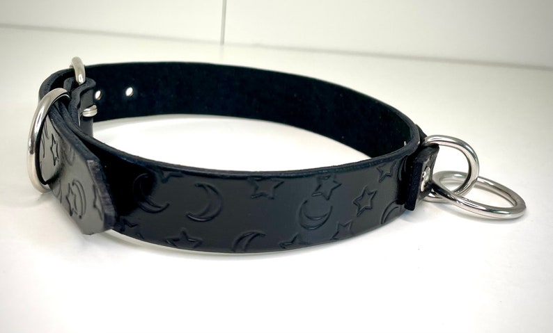 May include: Black leather collar with a moon and star pattern embossed on the surface. The collar has two silver D-rings.