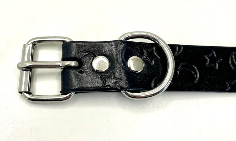 May include: Black leather dog collar with a silver buckle and D-ring. The collar has a moon and star pattern embossed into the leather.