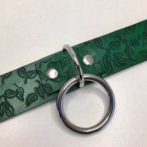 K&ouml;nnte beinhalten: Ein gr&uuml;nes Lederhalsband mit einem silbernen Metallring und zwei silbernen Metallnieten. Das Leder hat ein gepr&auml;gtes Blattmotiv.
