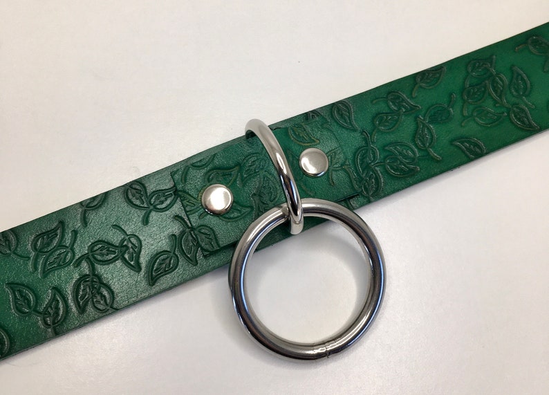 K&ouml;nnte beinhalten: Ein gr&uuml;nes Lederhalsband mit einem silbernen Metallring und zwei silbernen Metallnieten. Das Leder hat ein gepr&auml;gtes Blattmotiv.
