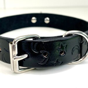 May include: Black leather dog collar with a silver buckle and D-ring. The collar is embossed with a repeating pattern of stars and crescent moons.