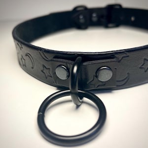 May include: Black leather choker with a star and crescent moon design. The choker has a black metal ring attached.