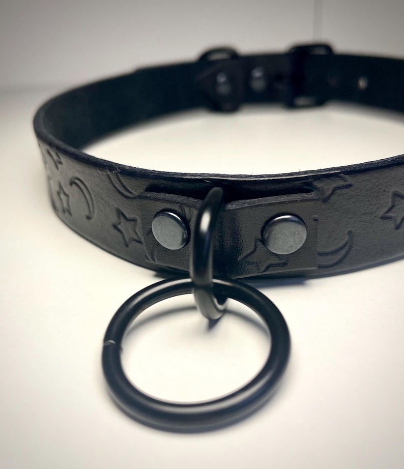 May include: Black leather choker with a star and crescent moon design. The choker has a black metal ring attached.
