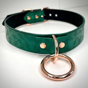 K&ouml;nnte beinhalten: Ein gr&uuml;nes Lederhalsband mit einem gepr&auml;gten Blattmotiv in Ros&eacute;gold und einem daran befestigten Ros&eacute;goldring.
